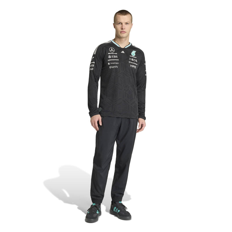adidas male model stands against a white studio background wearing a black Mercedes-AMG F1 long-sleeve training top and matching black trousers, with visible Mercedes and sponsor logos on the clothing.