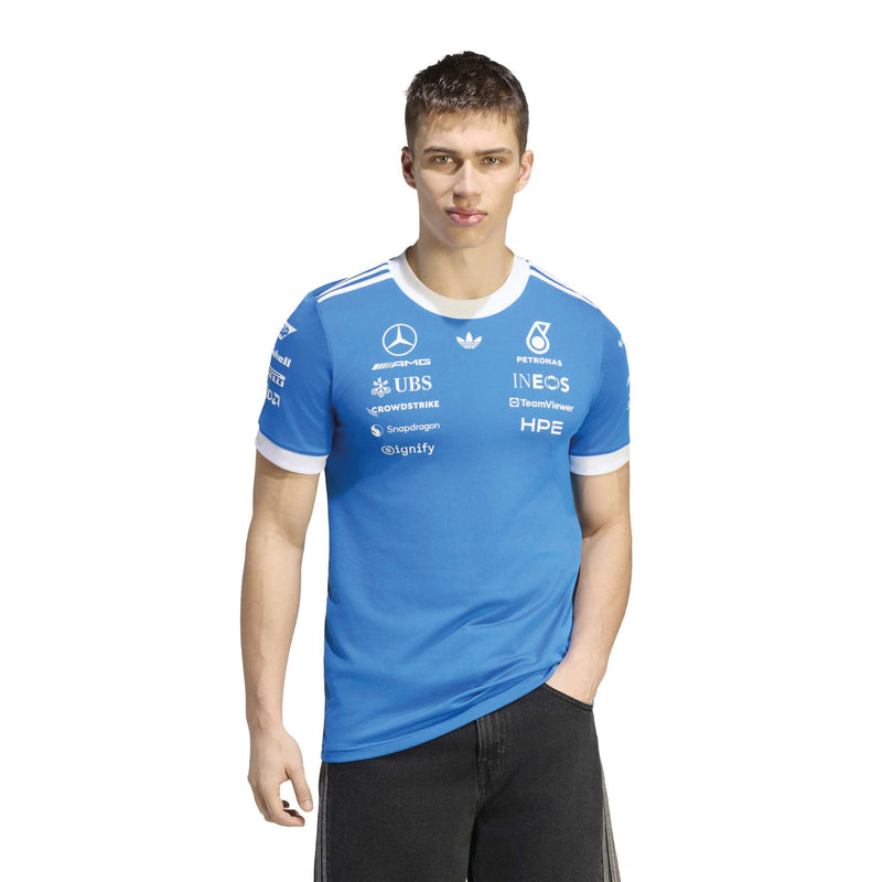 adidas man modeling a blue Mercedes Formula 1 team t-shirt with white sponsor logos and white trim on the collar and cuffs, paired with black pants, against a plain white background.